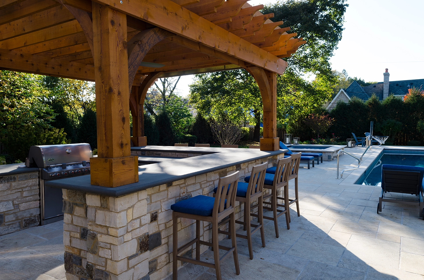 Terrace Design Construction outdoor kitchen under wooden pergola with stone masonry, stainless grill and full stone patio space surrounding pool